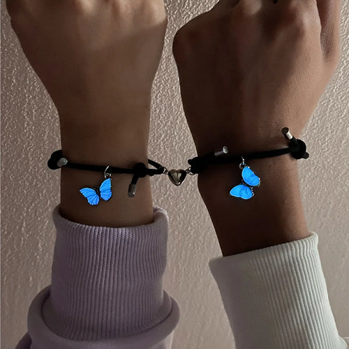 Two wrists wearing black bracelets with blue butterfly charms against a neutral background