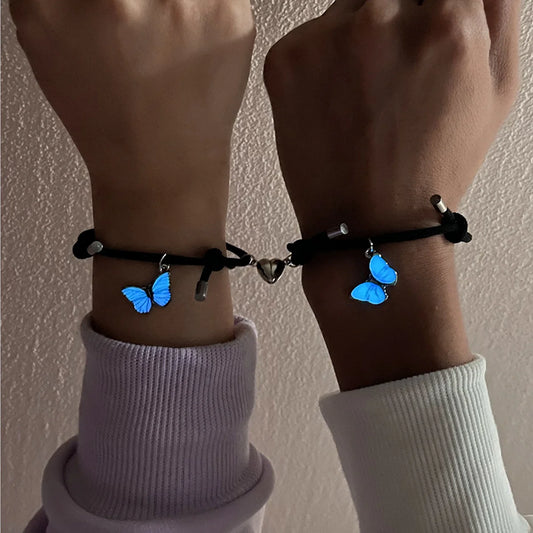 Two wrists wearing black bracelets with blue butterfly charms against a neutral background